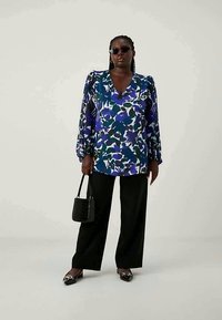 Blusa a fiori nei toni del blu e del verde, con maniche a sbuffo, scollatura a V, abbinata a pantaloni neri a gamba larga e una borsa nera texturizzata.