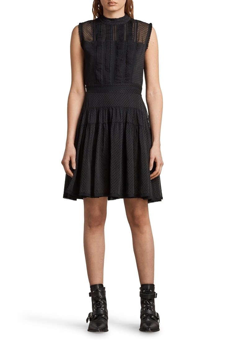 Femme portant une robe noire en dentelle sans manches avec des pois texturés et des bottines noires à boucles, debout devant un fond blanc.