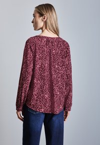 Blouse longline bordeaux avec un motif abstrait texturé, dotée d'un col froncé et d'une silhouette décontractée, associée à un jean sombre.