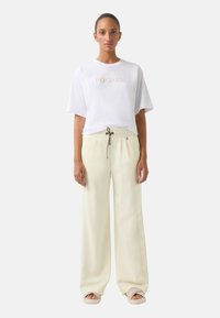T-shirt blanche avec le logo "BOGNER", accompagnée d'un pantalon large jaune clair avec une taille à cordon et des sandales beiges.