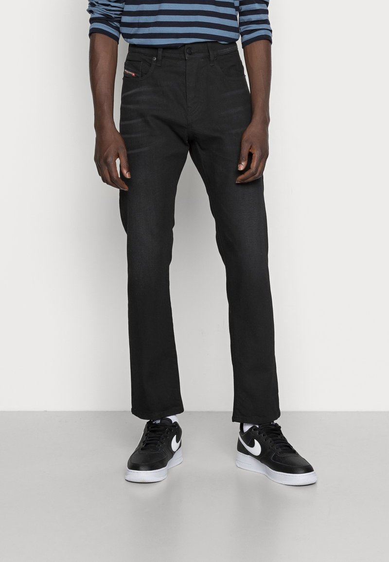 Homme portant un jean noir à jambes droites, des baskets Nike noires et blanches, et un t-shirt à manches longues rayé bleu et noir, debout devant un fond blanc.