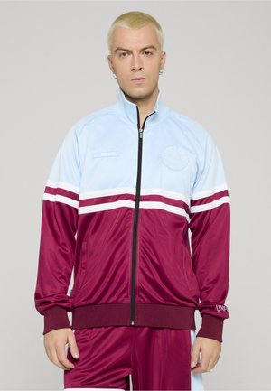 Junger Mann mit kurzen blonden Haaren trägt eine hellblaue und burgunderrote Sportjacke mit Reißverschluss und passende Hose, steht vor einfarbigem Hintergrund.