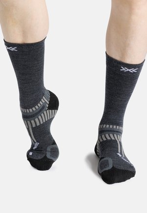 Pieds portant des chaussettes de sport gris foncé avec des motifs gris clair et noir, visibles de face et de dos sur fond blanc.