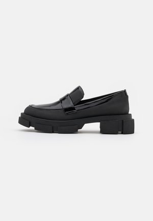 Sort loafers med chunky sål, glat finish og rem-detalje på toppen, vist fra siden på en ensfarvet lys baggrund.