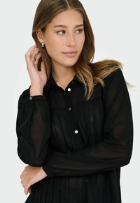 Černá, průhledná blůza s knoflíkovým zapínáním vpředu, sklady a dlouhými rukávy. Má klasický límec a kulaté knoflíky.