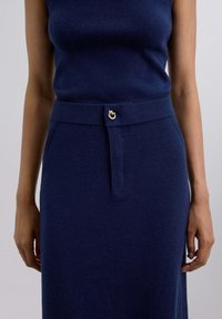 Close-up van een persoon die een marineblauw geribbeld mouwloos topje en een bijpassende geribbelde taille rok met een gouden ronde sluiting draagt.