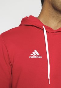 Sweat à capuche Adidas rouge avec logo blanc sur la poitrine et cordon blanc, porté par une personne cadrée au niveau du cou et de l'épaule.