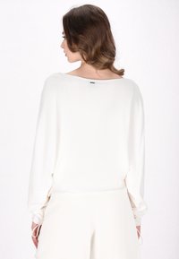 Top bianco, dalla vestibilità ampia, con scollatura larga e maniche lunghe; tessuto testurizzato, finitura opaca delicata, dettagli arricciati vicino ai polsi.