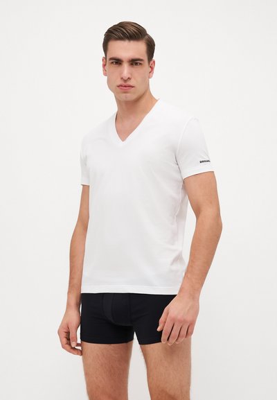 T-shirt blanc en tissu doux, à col en V, à manches courtes, avec un détail de logo noir sur le bras gauche, associé à un sous-vêtement noir ajusté.