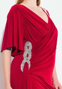 Červené šaty s krátkými rukávy, ozdobené jasnými rhinestones na boku. Hladká textura, hluboký výstřih a nařasený pas.