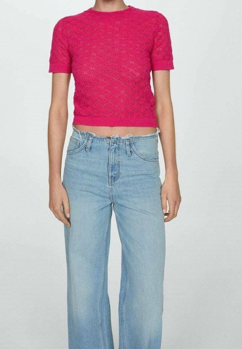 Personne portant un crop top en maille rose vif à manches courtes associé à un jean large taille haute de couleur bleu clair, sur un fond uni.