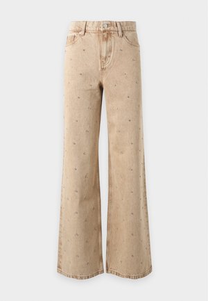 Kvinde iført en brun oversized frakke med sorte spænder, grå top, beige vidde bukser med små nitter og sorte loafers på hvid baggrund.