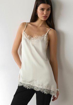 Young woman with long dark hair wearing a white lace-trimmed camisole top and black pants, standing against a white wall.