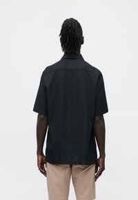 Camiseta preta de manga curta feita de tecido leve; apresenta uma gola apontada e costas direitas com detalhe em costura. Usada com calças de cor clara.