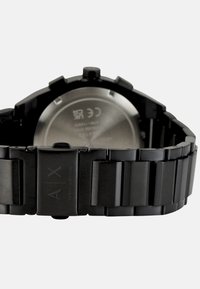 Reloj de pulsera negro de acero inoxidable con una correa de eslabones, que presenta un logo grabado y una tapa trasera plateada con un diseño minimalista.