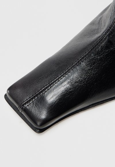 Czarny skórzany botek za kostkę z kwadratowym noskiem, gładką fakturą i przeszyciami wzdłuż boku. Podeszwa jest cienka i płaska.