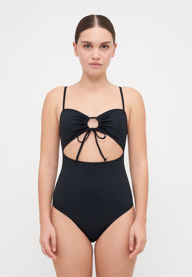 Maillot de bain une pièce noir avec des bretelles fines, une section médiane découpée, des détails froncés et un accent en anneau central. Texture de tissu lisse.