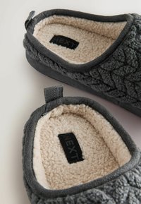 Šedé pantofle s texturovaným svrškem, měkkou krémovou podšívkou a smyčkovým poutkem pro snadné obouvání. Na stélce se nachází značka.