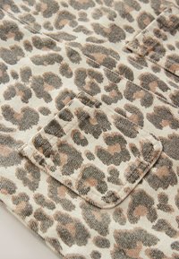 Tissu imprimé léopard avec un fond beige, présentant des taches noires et marron. Comprend un détail de poche cousu dans le design.