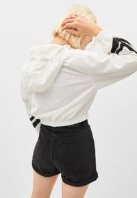 Biała kurtka przeciwwiatrowa z kapturem, z elastycznymi mankietami i dołem, z czarnymi paskami na rękawach. W zestawie z wysokimi czarnymi szortami dżinsowymi.