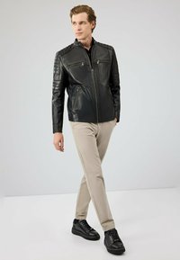 Zwarte leren jas met gewatteerde schouderdetails, twee ritszakken aan de voorkant, gecombineerd met beige chinos en zwarte sneakers. Minimalistisch ontwerp.