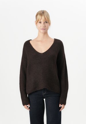 Kvinne med blondt hår iført en løs, mørkebrun V-hals genser og mørke jeans, som står mot en vanlig hvit bakgrunn.