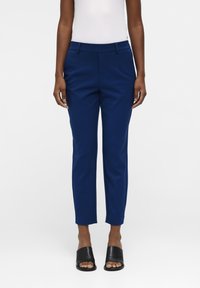Persoon in slim-fit marineblauwe broek, witte mouwloze top en zwarte open teen instapschoenen, staande tegen een effen achtergrond.