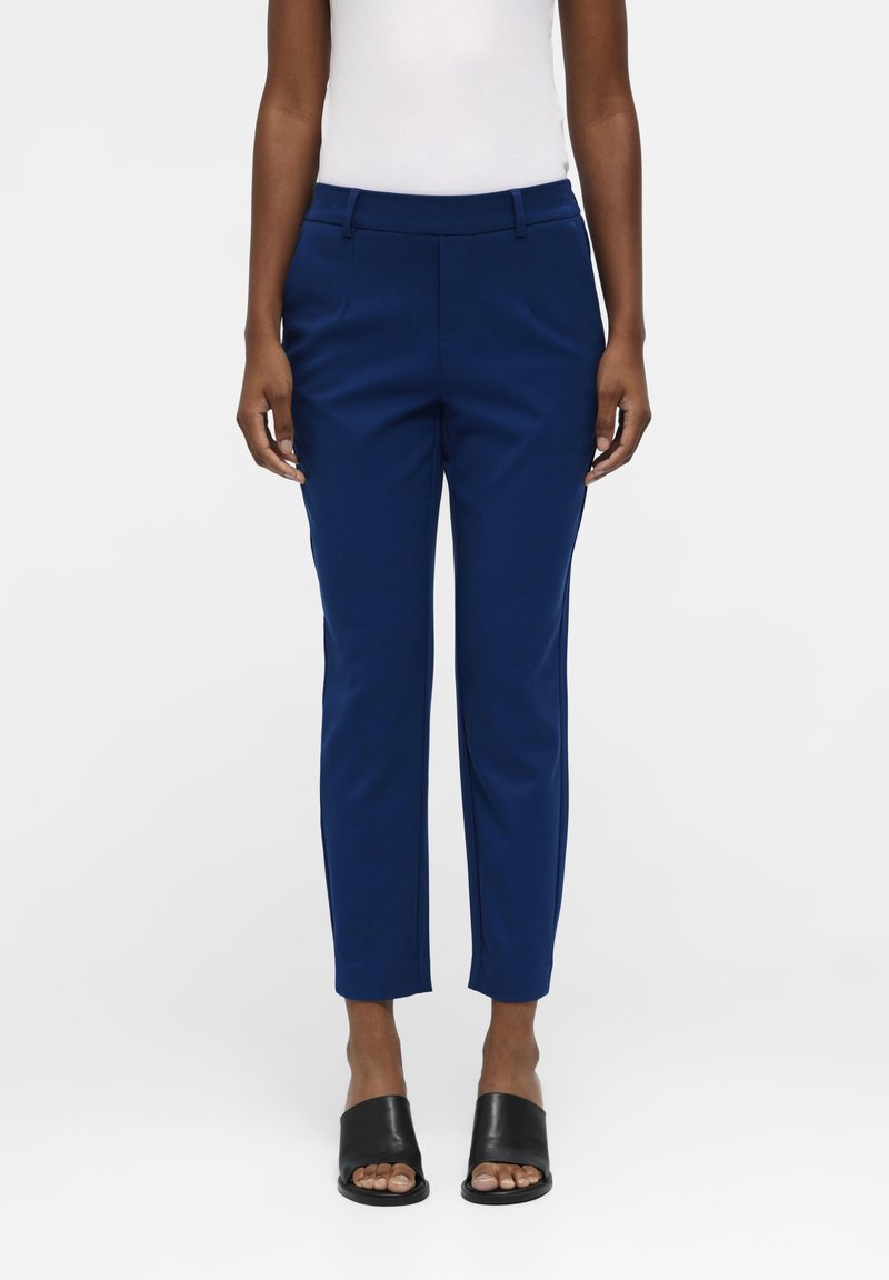 Persoon in slim-fit marineblauwe broek, witte mouwloze top en zwarte open teen instapschoenen, staande tegen een effen achtergrond.