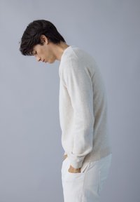 Maglione a maglia beige chiaro con scollatura rotonda e polsini a coste, abbinato a pantaloni bianchi. Il tessuto sembra morbido e accogliente.