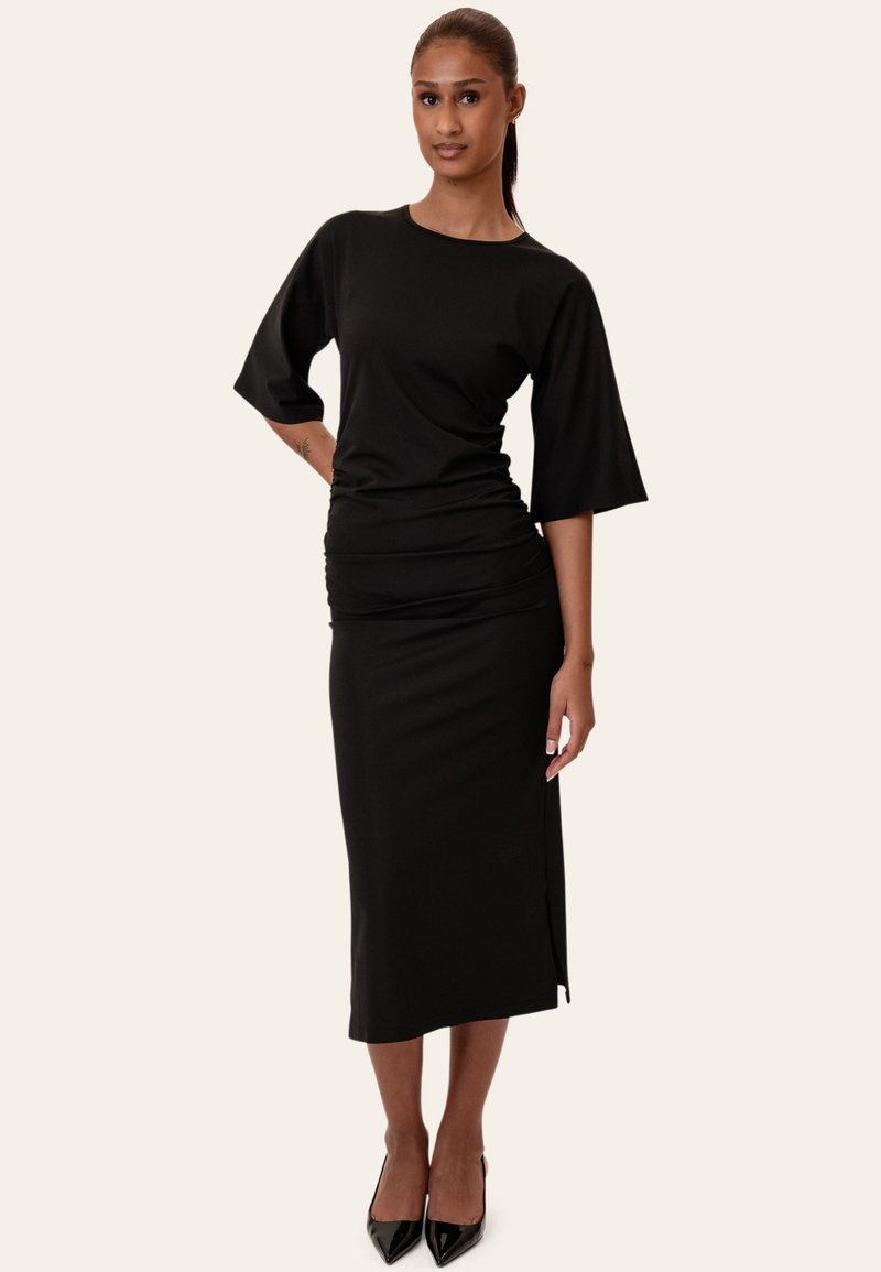 Femme debout, main sur la hanche, portant une robe midi noire à manches aux coudes et des talons hauts noirs sur un fond uni.
