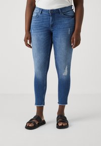 Jeans ajustados azules en material denim con detalles desgastados, un pequeño desgarro en una rodilla y dobladillos deshilachados hasta el tobillo, combinados con sandalias negras de tipo deslizador.