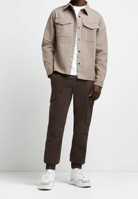 Beżowa koszula zapinana na guziki z dwoma kieszeniami na piersi, zestawiona z ciemnobrązowymi spodniami typu cargo z bocznymi kieszeniami. Białe trampki z szarawymi akcentami.