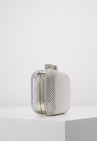 Petit sac à main rectangulaire en argent avec des rhinestones et un fermoir orné, posé sur un piédestal blanc contre un fond uni.