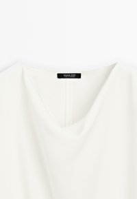 Hvid V-hals bluse med en glat tekstur, der har en subtil glans og en bagsømsdetalje. Etiketten læser "Massimo Dutti, Fremstillet i Marokko."