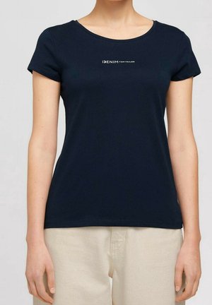 Kobieta nosząca dopasowany granatowy T-shirt z napisem „DENIM TOM TAILOR” na piersi, zestawiony z jasnobeżowymi spodniami.