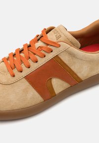 Sneaker beige in suede con lacci e dettagli arancioni. Presenta un pannello laterale marrone testurizzato e una suola in gomma con una finitura liscia.