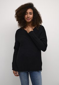 Schwarzer gerippter Pullover mit V-Ausschnitt und langen Ärmeln. Hat eine lockere Passform und Seitenschlitze und wird mit Jeans kombiniert.