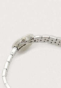 Sudraba metāla pulksteņa josta ar saistītu aproci un apaļu pulksteņa korpusu, skatīta no sānu, uz balta fona redzams pulksteņa kronis.