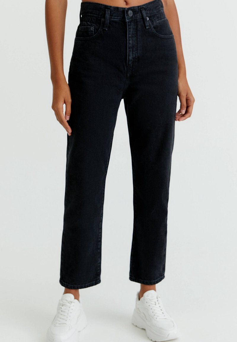 Femme portant un jean noir taille haute à jambes droites et des baskets blanches épaisses, debout devant un fond clair uni.