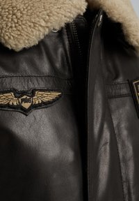 Chaqueta de cuero negro con un collar de piel de oveja color beige, emblema de alas bordado y cierre de cremallera diagonal. Textura suave y diseño duradero.