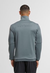 Szara, ocieplana kurtka zapinana na zamek z wysokim kołnierzem, ściągaczami przy rękawach oraz wytłoczonym logo "Sergio Tacchini" na plecach. Gładki, średniej wagi materiał.