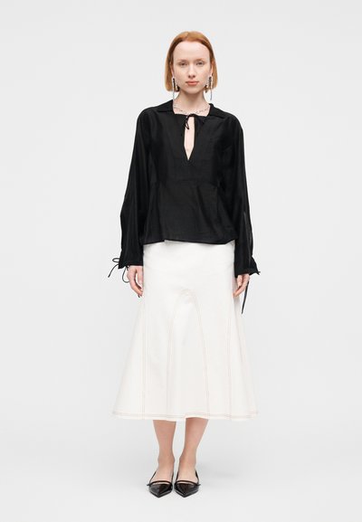 Blouse noire à manches longues avec un col en V, des poignets plissés et un détail noué. Jupe blanche à forme évasée avec des coutures contrastantes. Chaussures plates noires à bout pointu.