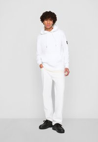 Felpa bianca con cappuccio e stringhe, logo sulla manica, abbinata a pantaloni bianchi e scarpe nere. Tessuto morbido, vestibilità rilassata, design moderno.