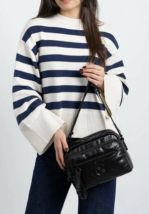 Bolso cruzado de cuero negro con un bolsillo con cremallera y acentos texturizados, sostenido por una persona que lleva un suéter a rayas azul marino y blanco.