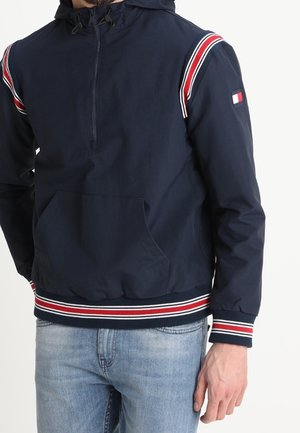 Mand iført navyblå hættetrøje med røde, hvide og blå striber på ærmer, kant og manchetter, kombineret med lyseblå jeans.