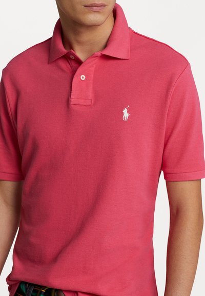 Polo à manches courtes en coton rose vif, avec un col classique, une patte de boutonnage à trois boutons et un logo brodé blanc sur la poitrine.