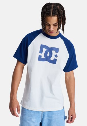 Giovane uomo con i capelli intrecciati, che indossa una T-shirt bianca e blu navy con il logo DC e jeans azzurri, guarda verso il basso.
