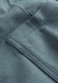 Prim-plan al unei țesături texturate de culoare teal, cu un buzunar mic dreptunghiular și cusături vizibile.