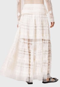 Λευκή φούστα με στρώσεις δαντέλας και βολάν στο τελείωμα, με διάφανα τμήματα και λεπτομερείς δαντελένιες λεπτομέρειες. Φορεμένη με μαύρα σανδάλια επίπεδου πάτου.