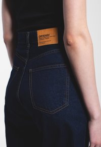 Tmavě modré džíny mají vysoký pas, oranžové švy a na zadní straně obdélníkovou koženou nášivku označenou "DRDENIM".